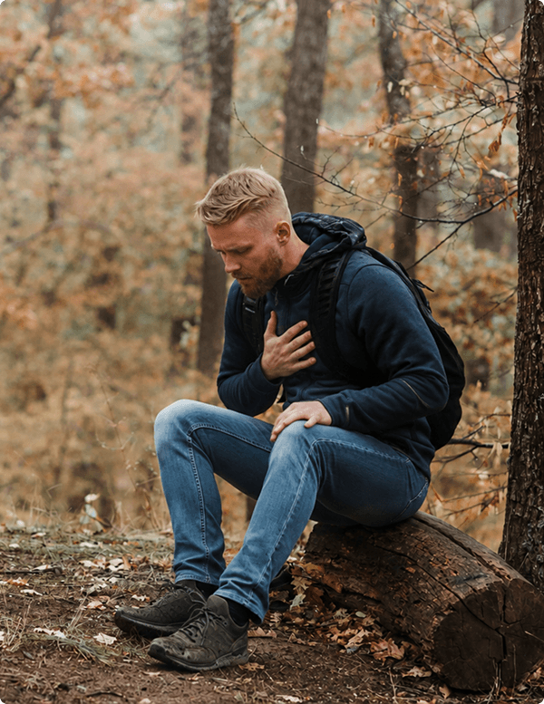 A man with blond hair sits on a log in a forest, clutching his chest with a concerned expression. He's wearing a navy hoodie and jeans. The autumn setting conveys a tense mood.